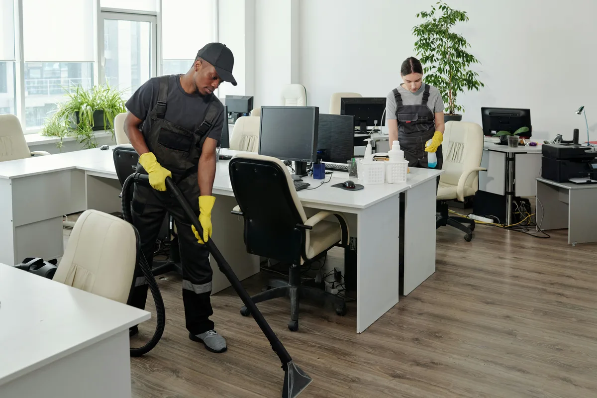 Person vacuuming an office floor while another wipes a desk