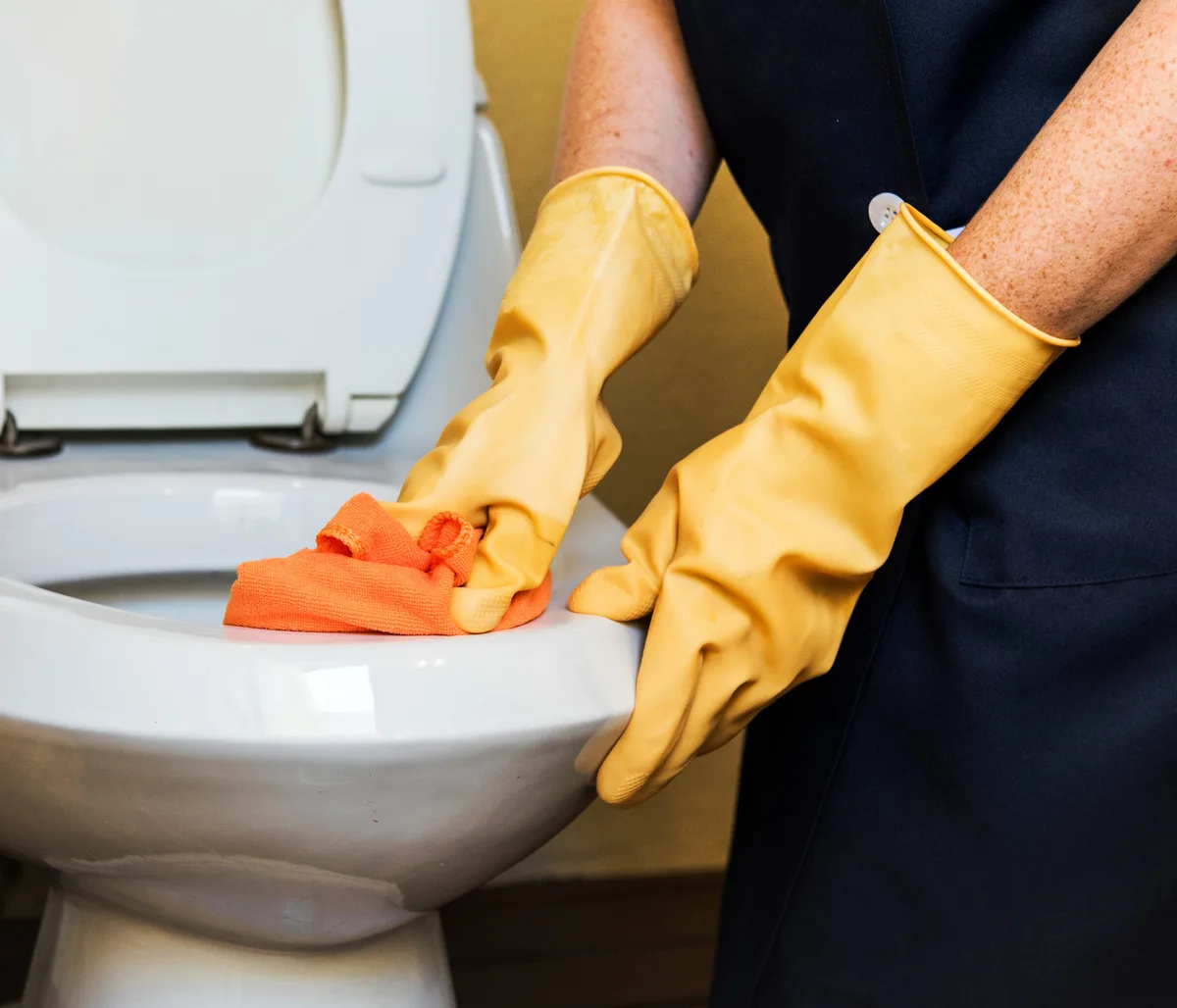 Gloved hands wiping the outside of a toilet with a cleaning cloth